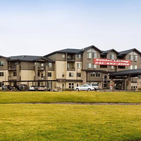 Exterior view of a multi-story senior living facility building with a covered entrance and several parked cars in front. The building has beige and brown siding with stone accents and balconies. A large red banner on the building reads 'Senior Living 541-497-2106'.