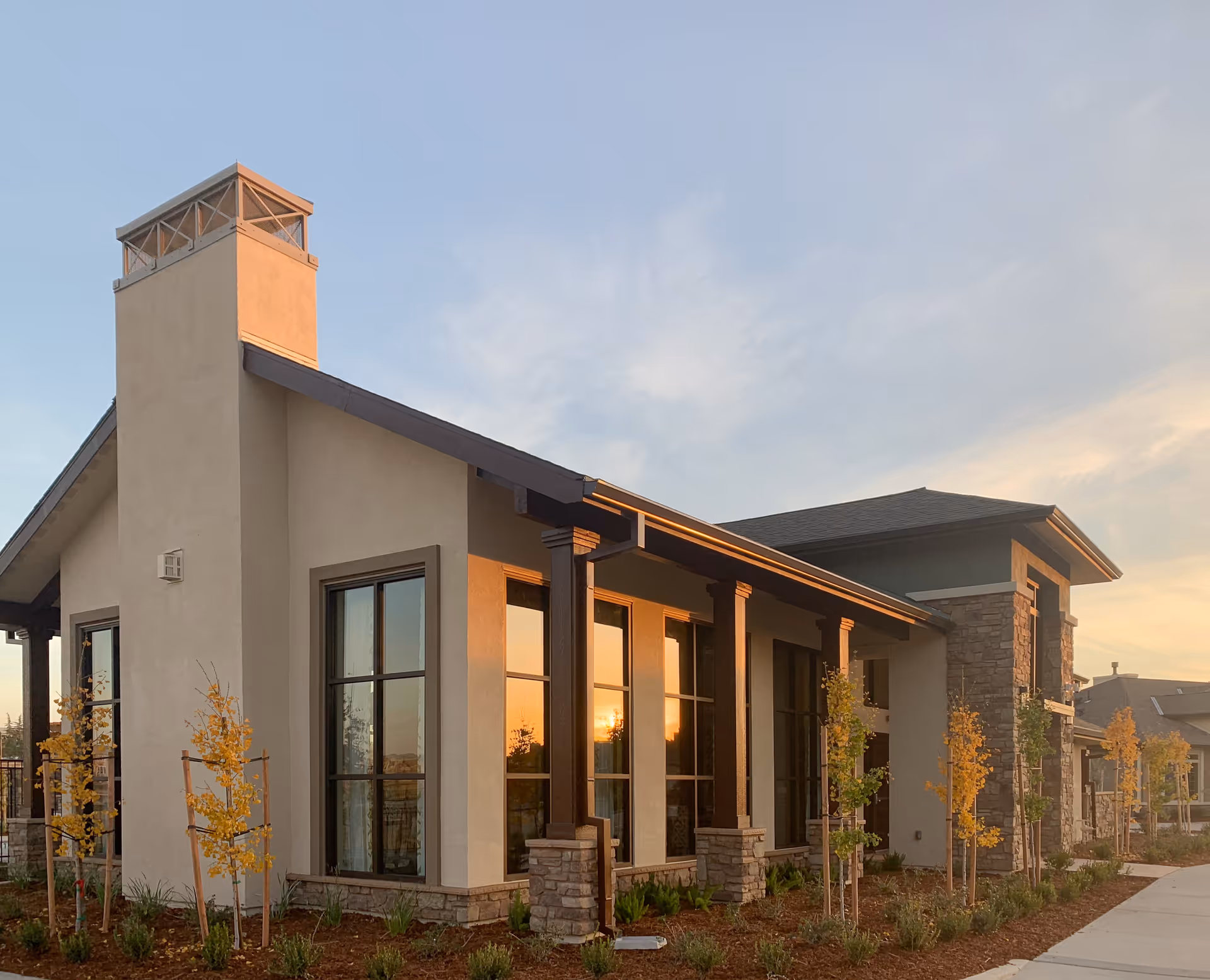 Exterior front of a modern single-story senior living facility with large windows, columns, and young landscaping at sunset.