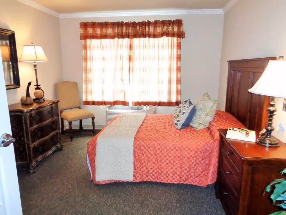 A cozy bedroom with a bed covered in a red quilt and decorative pillows. There is a wooden nightstand with a lamp and a book on it next to the bed. Across from the bed is a wooden dresser with decorative items and a chair beside it. A window with red and beige plaid curtains lets in natural light.