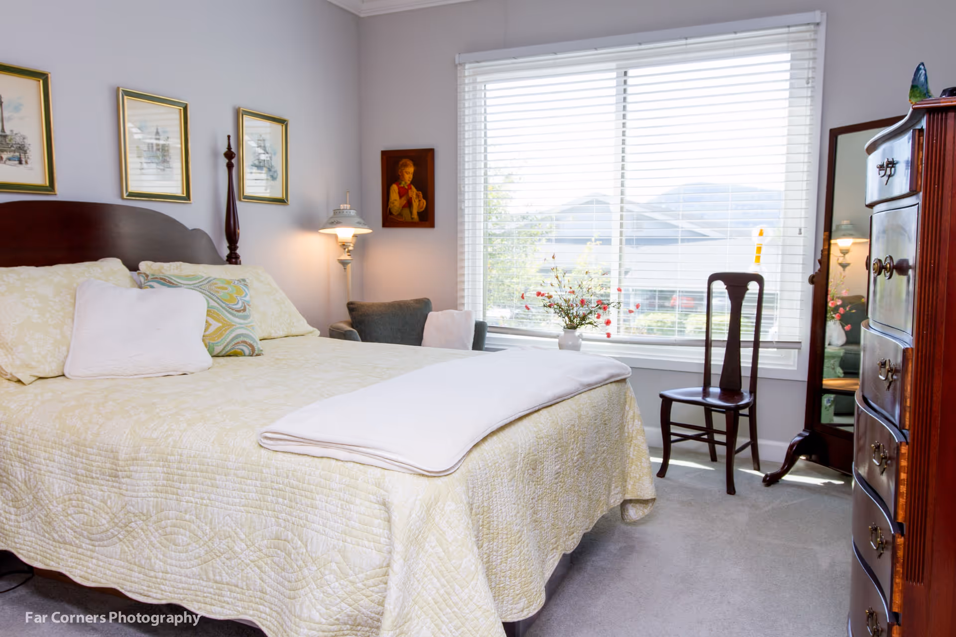 A bright bedroom with a large bed covered in a yellow quilt and multiple pillows. There is a wooden headboard, three framed pictures on the wall above the bed, and a vintage-style lamp beside an armchair in the corner. A window with white blinds lets in natural light, and a wooden chair and dresser with a mirror are positioned near the window. A vase with flowers sits on the windowsill.