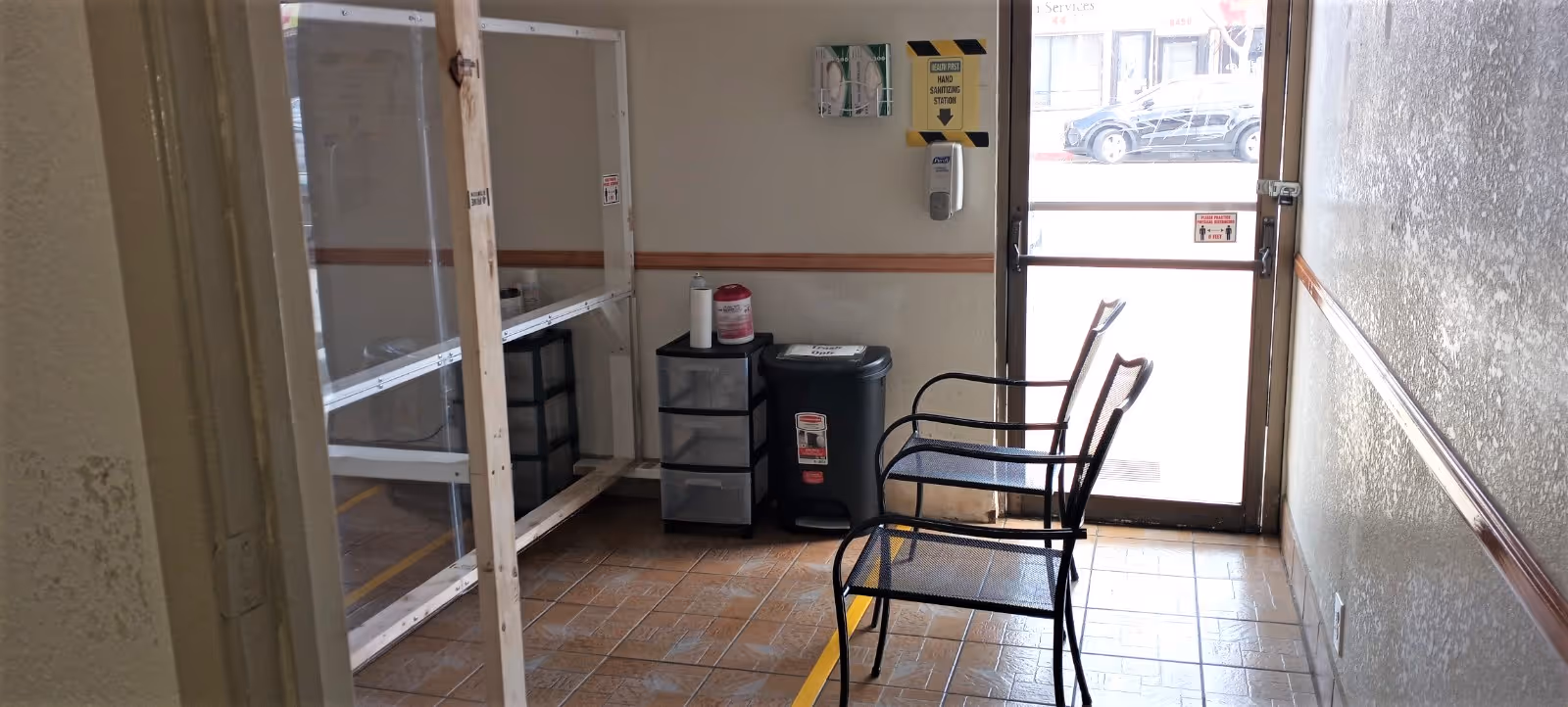 Small waiting area inside a facility with two black metal chairs, a trash bin, a small drawer unit, a hand sanitizer dispenser mounted on the wall, and a clear plastic barrier supported by wooden frames. The area has tiled flooring and a glass door leading outside where a parked car is visible.