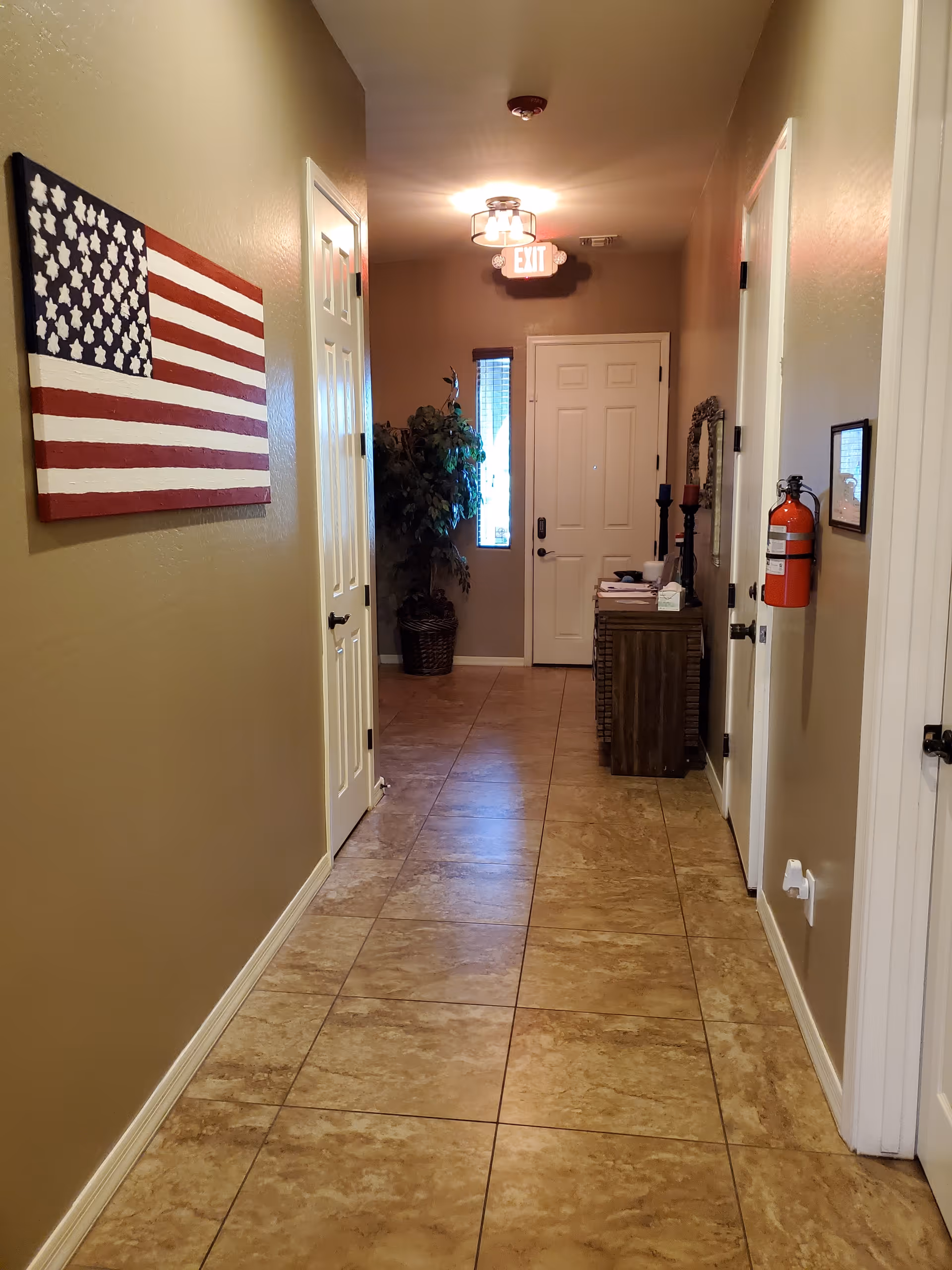Tile-floored hallway in an assisted living facility with an American flag wall art, exit sign, and a door at the end.
