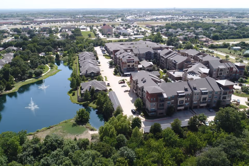 Aerial view of a senior living facility named Watercrest at Mansfield, showing multiple residential buildings with gray roofs surrounded by trees and greenery. There is a pond with two water fountains on the left side and parking areas between the buildings. The surrounding area includes more trees, roads, and distant buildings under a clear sky.