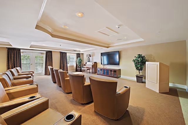 A bright communal media room with rows of leather recliners facing a large wall-mounted TV and a piano.
