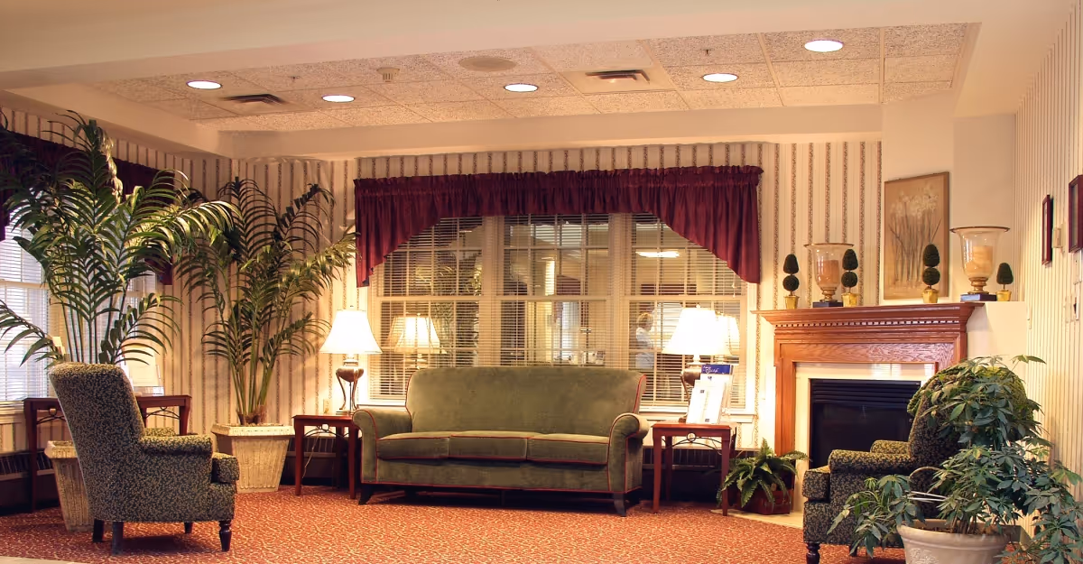 A cozy living room area with a green upholstered sofa centered between two wooden side tables with lamps. Two patterned armchairs face the sofa. There are large potted plants on both sides of the room, a fireplace with decorative items on the mantel, and a large window with blinds and a burgundy valance behind the sofa. The room has patterned wallpaper and a carpeted floor.