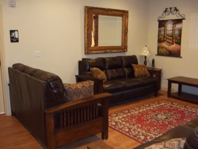 Small living room with dark leather sofas, wooden furniture, a patterned rug, and framed mirror and wall art.