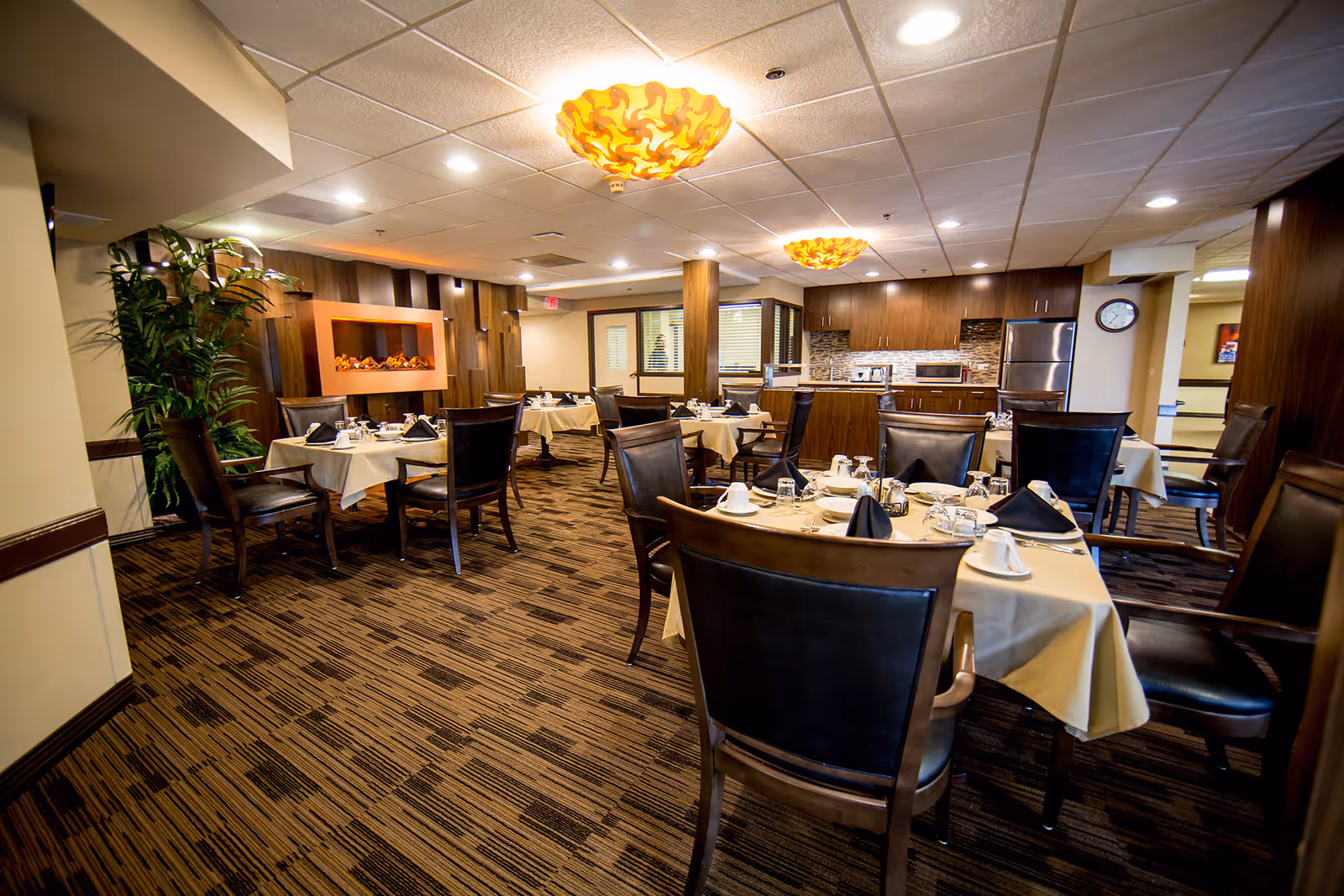 A dining room with several tables covered in beige tablecloths, each set with black napkins, cups, glasses, and silverware. The room features dark wooden chairs with black cushions, a patterned carpet, two decorative ceiling lights, a modern electric fireplace on the left wall, and a kitchen area with stainless steel appliances in the background.