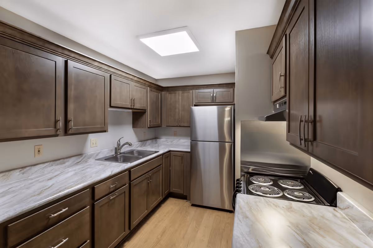 A modern kitchen with dark wooden cabinets, a stainless steel refrigerator, an electric stove with coil burners, a double sink, and marble-patterned countertops. The floor is light wood, and a bright ceiling light illuminates the space.