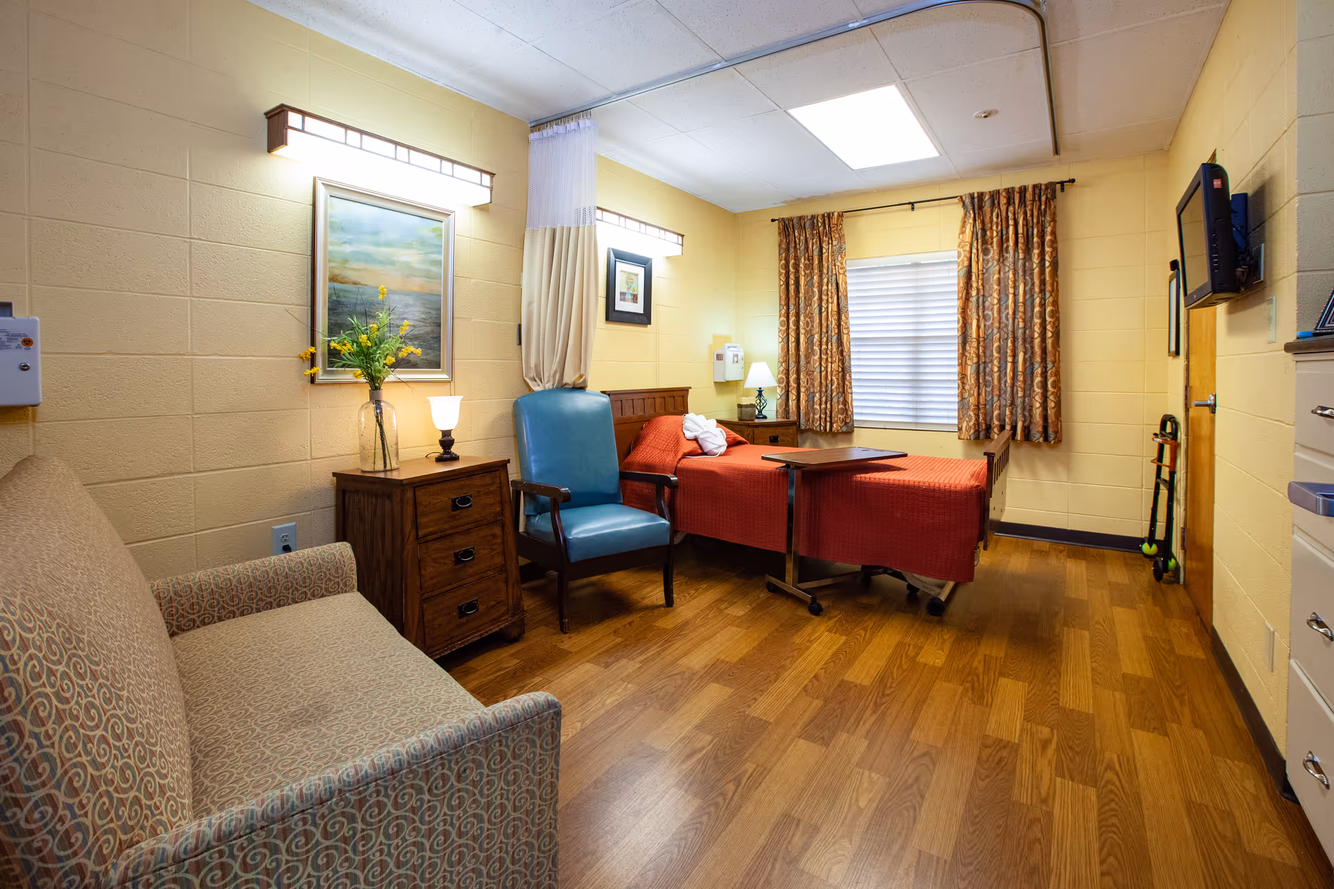 A cozy patient room in a senior living facility with a single bed covered in a red blanket, a blue armchair, a wooden nightstand with a lamp and a vase of yellow flowers, a patterned sofa, and a window with patterned curtains. The room has wood flooring and soft lighting fixtures on the walls.