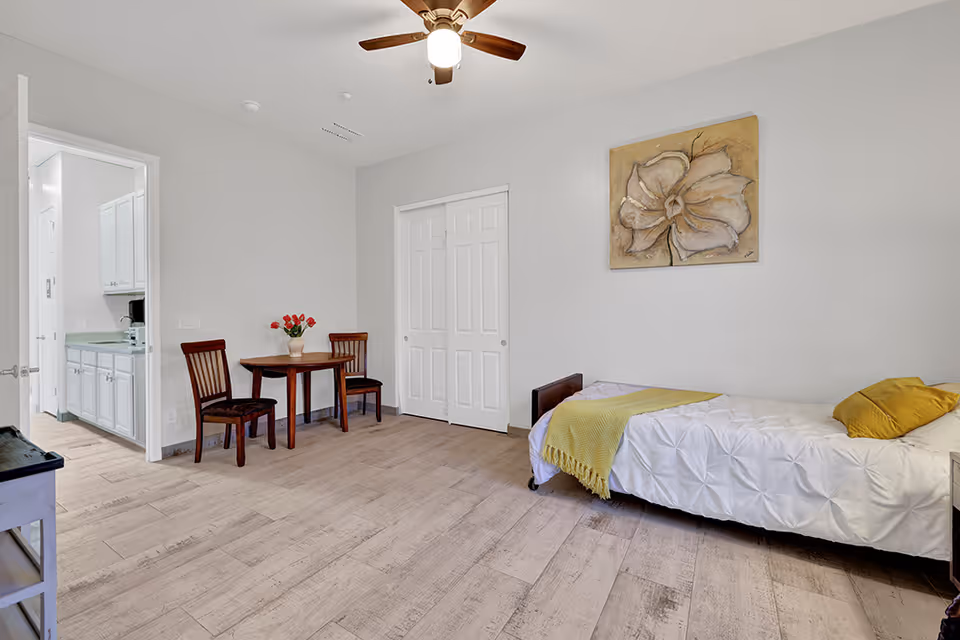 A bright bedroom with a single bed covered in a white textured bedspread and a yellow throw blanket and pillow. There is a wooden table with two chairs and a vase of red flowers near the open doorway leading to a kitchen area with white cabinets. A large floral painting hangs on the wall above the bed, and a ceiling fan with a light is mounted on the ceiling.