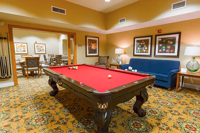 Interior view of a recreational room featuring a red felt pool table in the center. Behind the pool table is a blue couch with two framed artworks on the wall above it. There are two side tables with lamps on either side of the couch. In the background, there is an adjoining room with a dining table and chairs. The room has patterned carpet flooring and warm lighting.