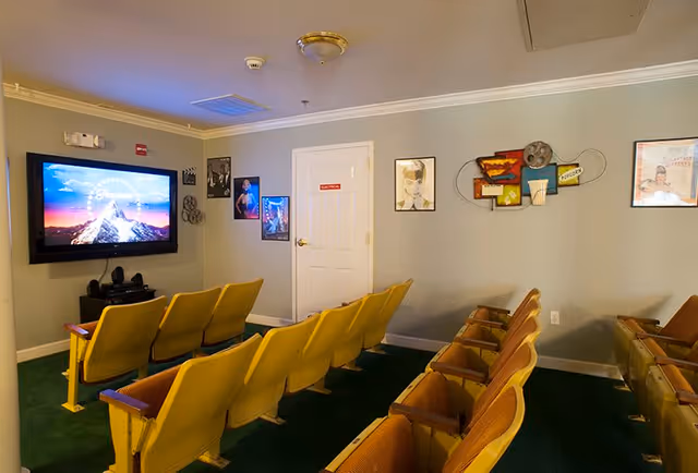 A small movie theater room with two rows of yellow theater seats facing a large flat-screen TV mounted on the wall. The walls are decorated with framed movie posters and film-themed decor. The room has a green carpet and a white door labeled 'Exit'.