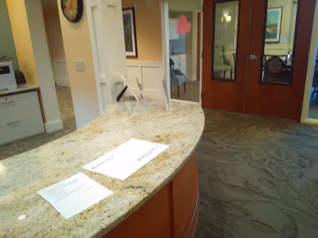 Interior view of a reception or front desk area with a curved granite countertop, papers and a clear acrylic sign holder on the counter, beige walls, carpeted floor, and double wooden doors with glass panels in the background.