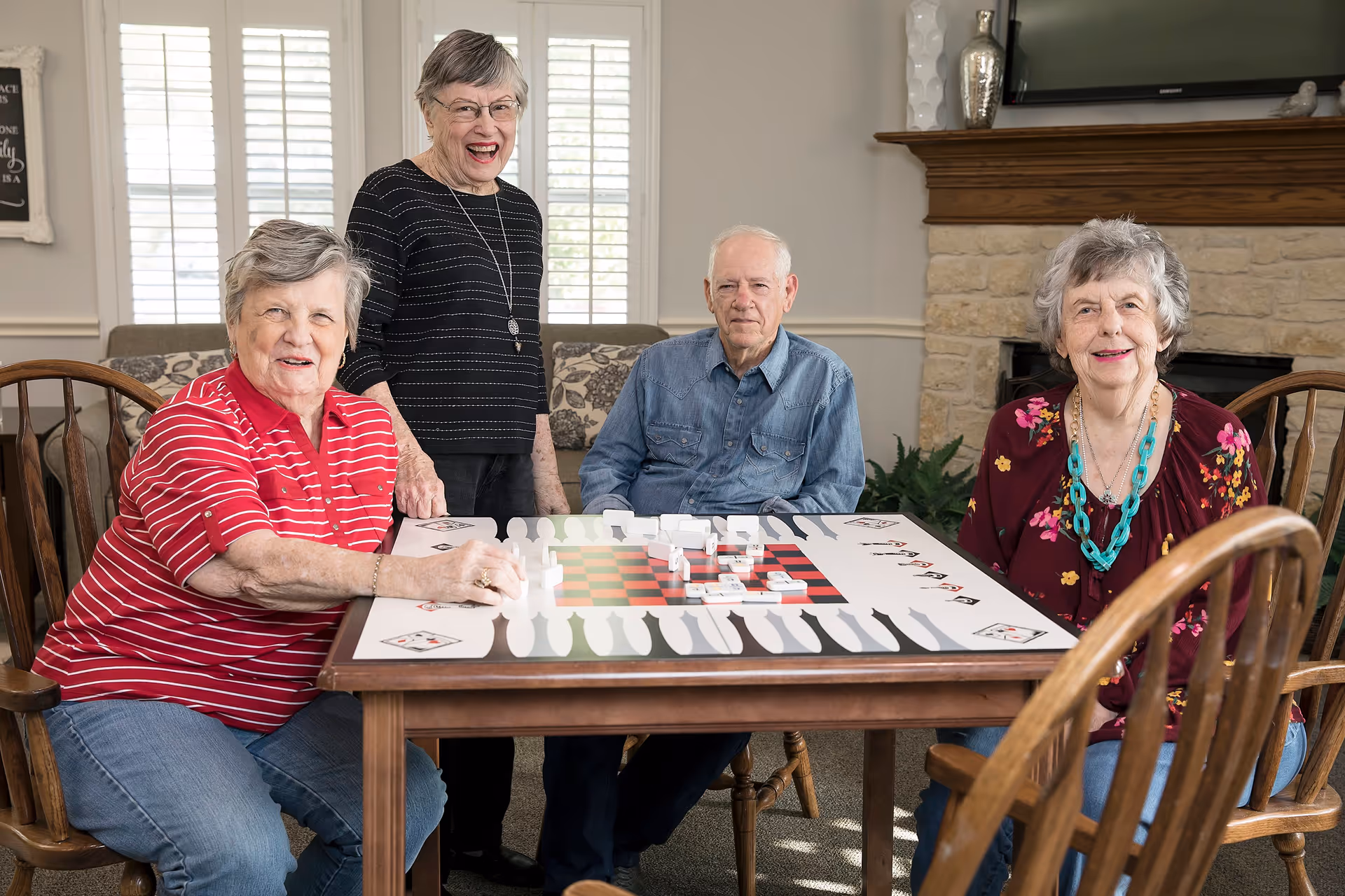 Four elderly individuals gathered around a table playing a large chess and checkers board game in a cozy living room with a stone fireplace and a TV mounted above it.