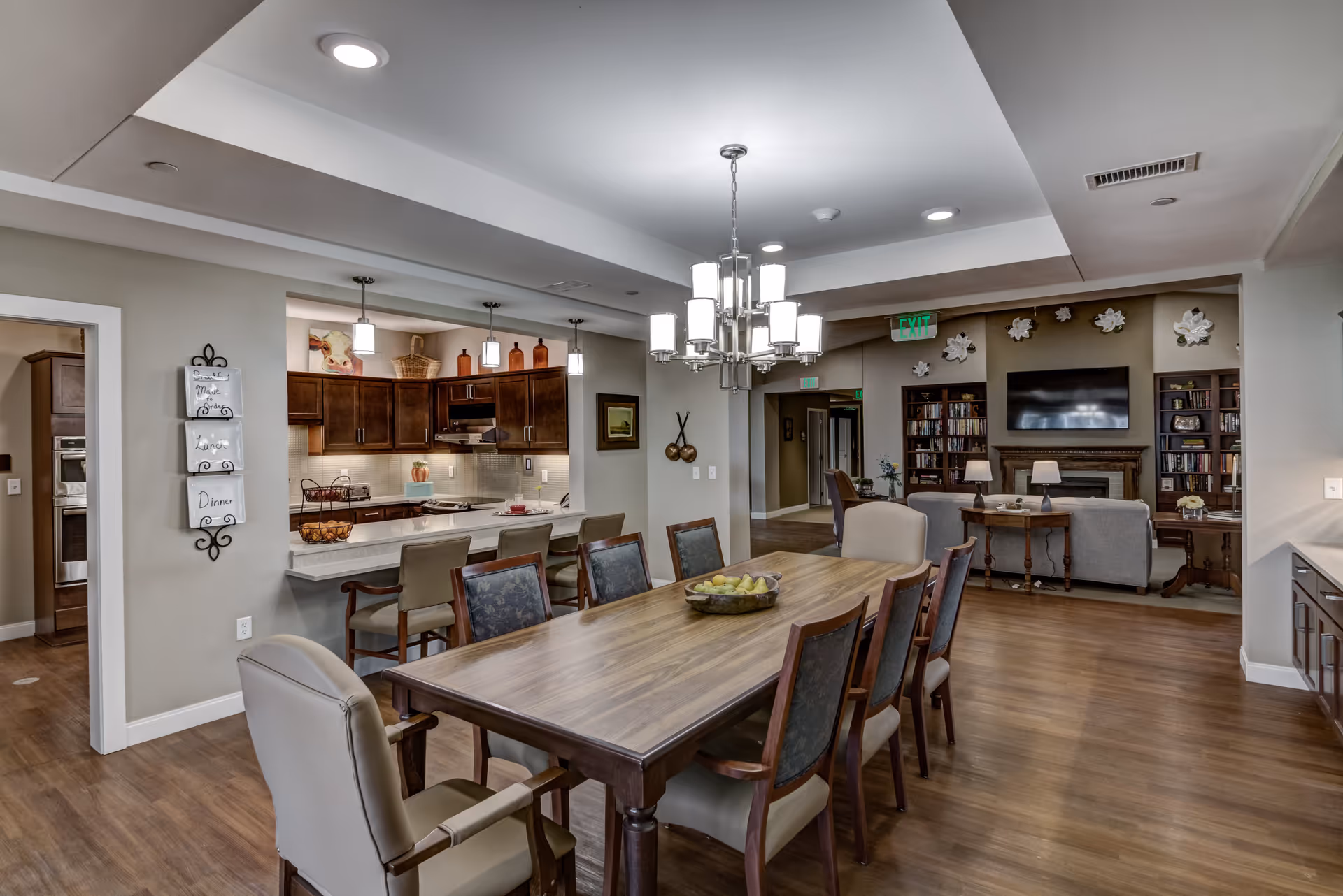 A spacious dining and living area in a senior living facility. The foreground features a large wooden dining table with eight chairs, a bowl of fruit on top, and a modern chandelier hanging above. To the left is a kitchen with dark wood cabinets, a countertop with bar stools, and pendant lights. In the background, there is a living room area with a fireplace, a wall-mounted TV, bookshelves, and decorative wall art. The flooring is wood, and the walls are painted in neutral tones.