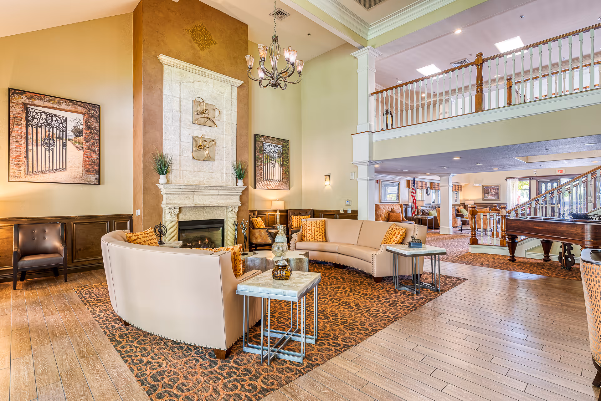 Spacious senior living lobby with curved sofas arranged around a fireplace, chandelier, and an open mezzanine balcony.