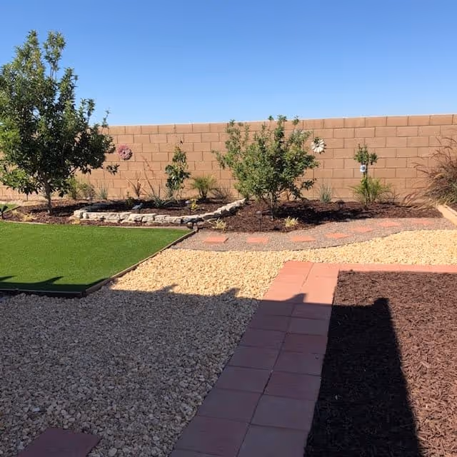Backyard garden with gravel paths, a brick walkway, artificial turf, and small trees and shrubs along a cinder block wall.