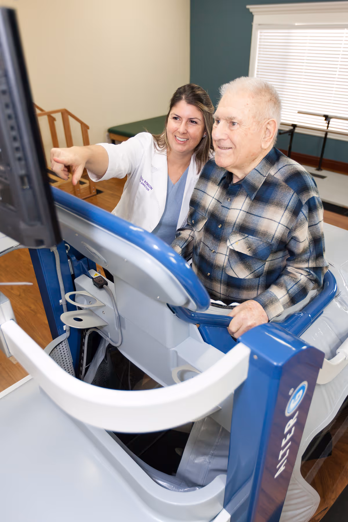 A healthcare professional in a white coat is assisting an elderly man who is seated on a blue and white exercise machine, pointing at a screen in front of them in a well-lit room with wooden flooring and a window with blinds.