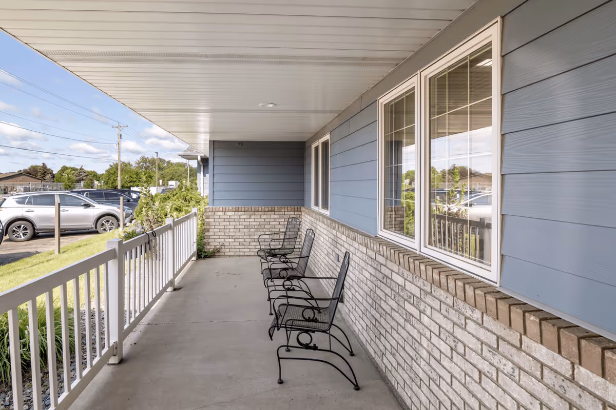 Covered outdoor patio area with three black metal chairs lined up against a brick and blue siding wall, overlooking a parking lot with several cars and greenery in the background.
