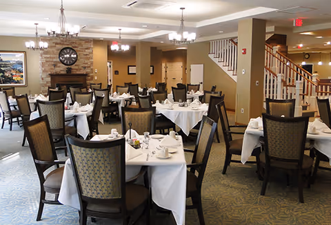 A spacious dining room with multiple round tables covered with white tablecloths, set with cups, saucers, and napkins. The room features cushioned chairs with patterned backs, a stone fireplace with a large clock above it, chandeliers hanging from the ceiling, and a staircase leading to an upper floor.