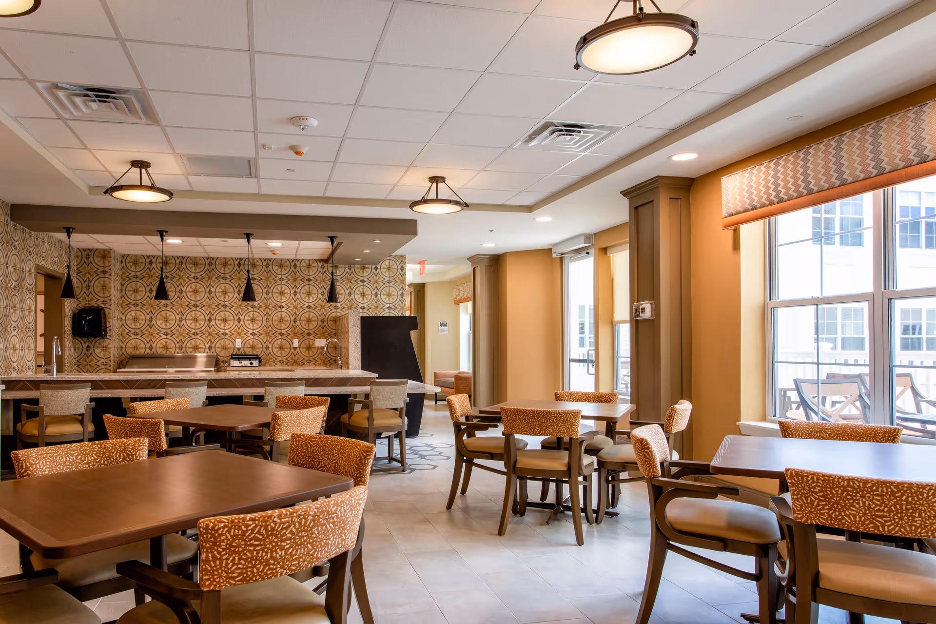 Bright dining room with multiple wooden tables and patterned chairs, a serving counter with pendant lights, and large windows letting in natural light.
