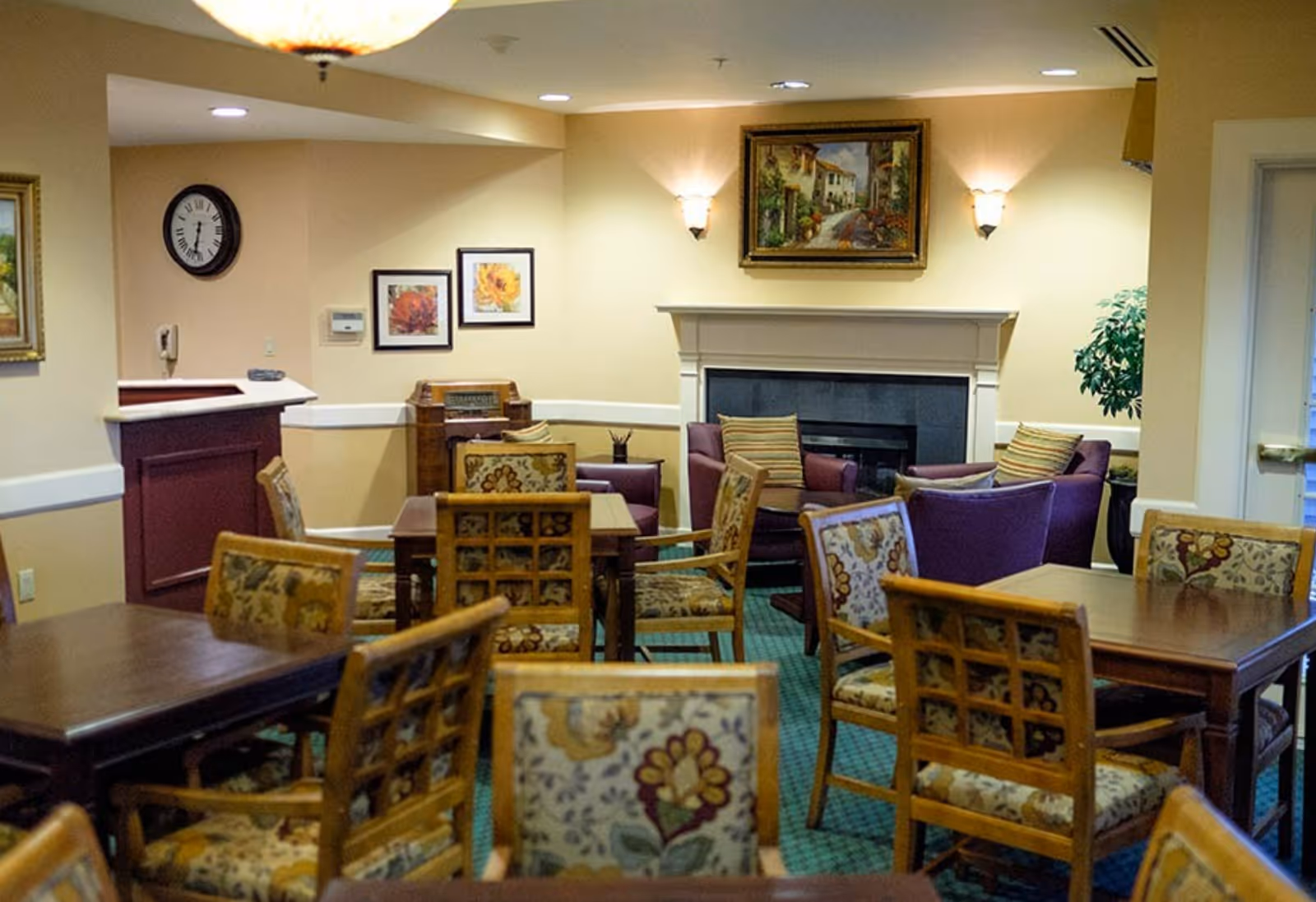 A cozy common area with multiple wooden tables and floral upholstered chairs arranged around the room. In the background, there is a fireplace with a framed painting above it, two purple armchairs with striped cushions, and wall sconces providing warm lighting. The walls are painted beige, and there are additional framed artworks and a clock on the walls.