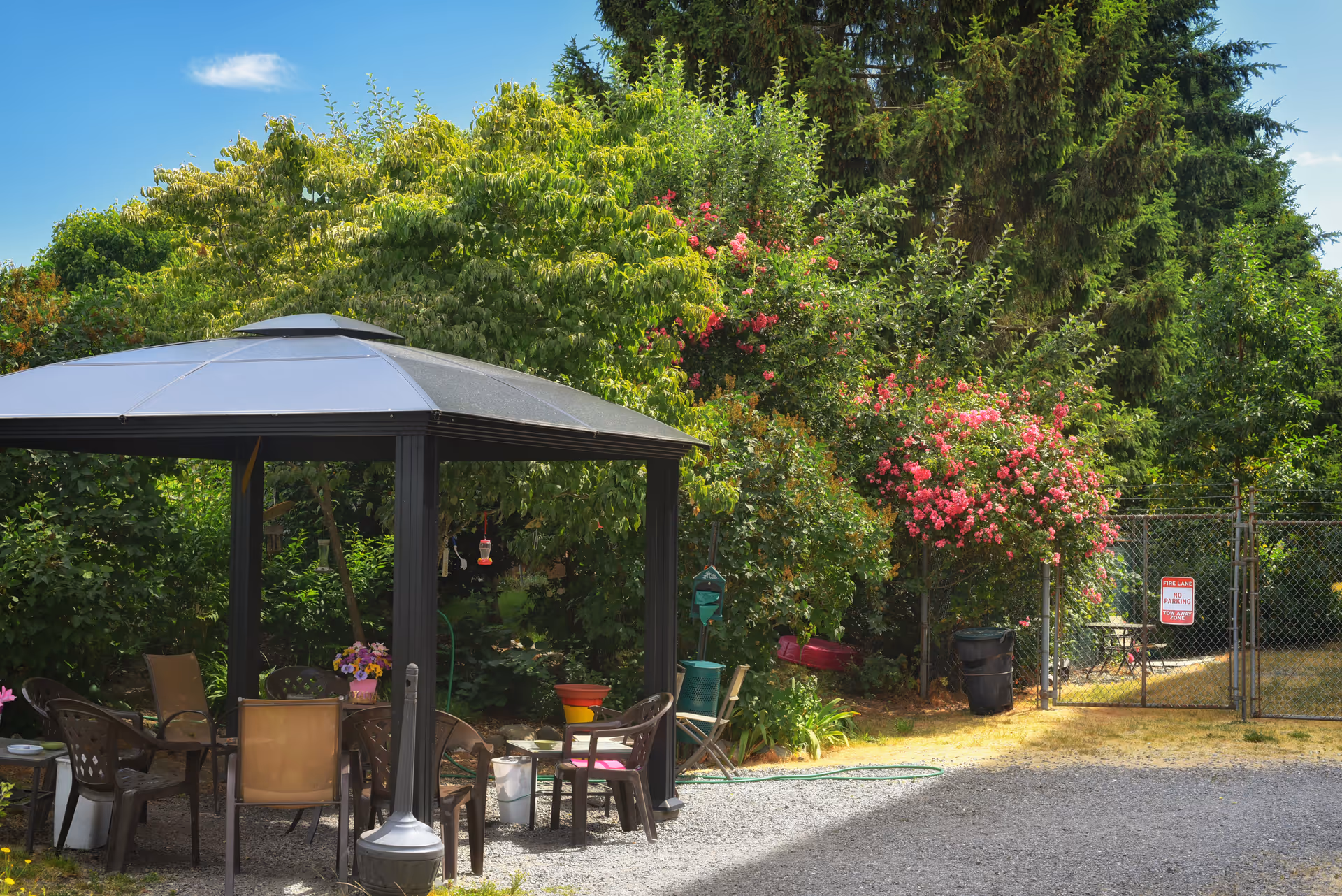 Outdoor garden area with a black metal gazebo covering several brown plastic chairs and a table with a flower pot on it. Surrounding the gazebo are lush green trees and bushes, including a flowering bush with pink blossoms. A chain-link fence with a gate and a 'Fire Lane No Parking' sign is visible in the background under a clear blue sky.