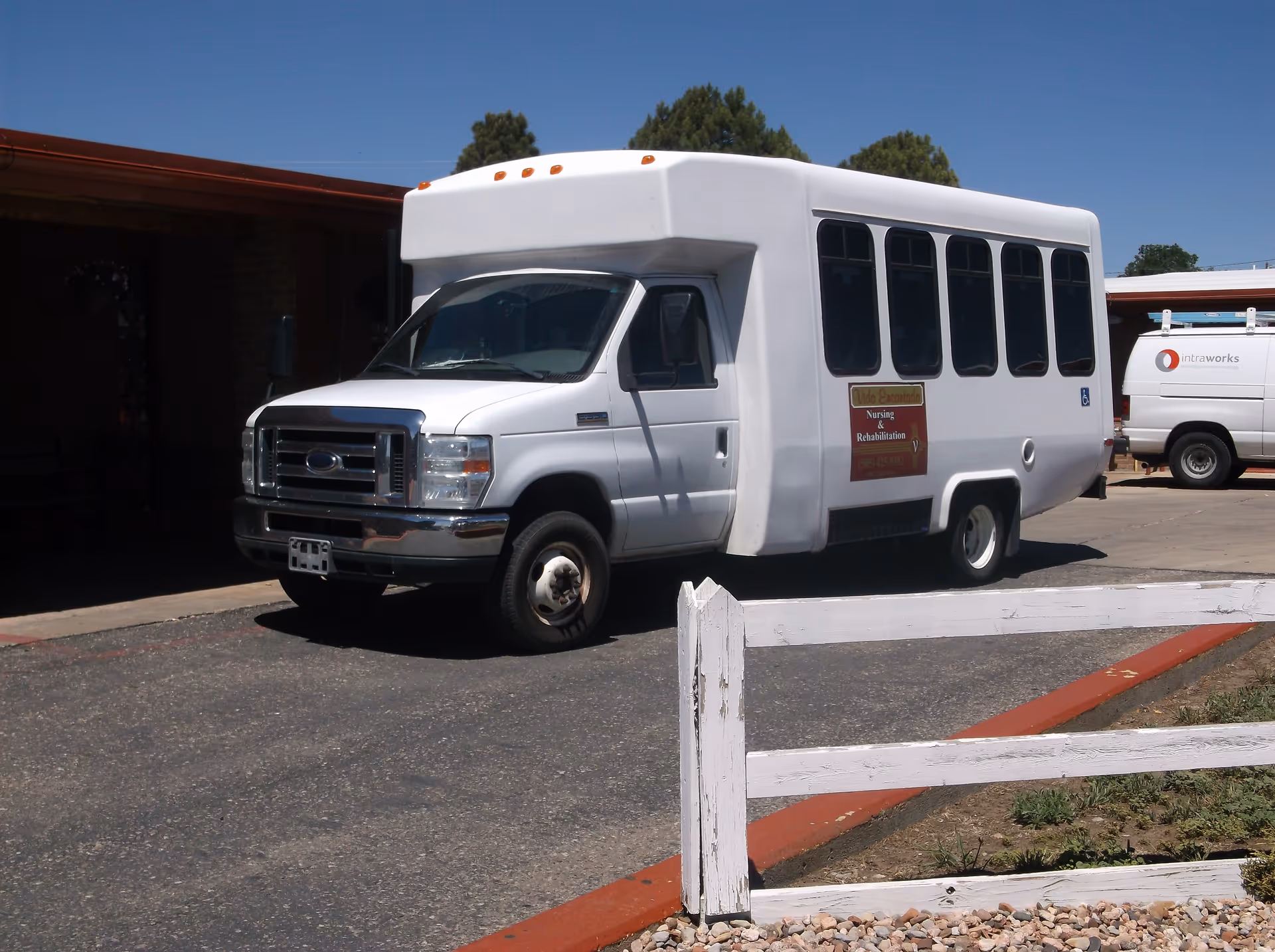 White passenger shuttle bus parked in front of a single-story building beside a white wooden fence under a clear blue sky.