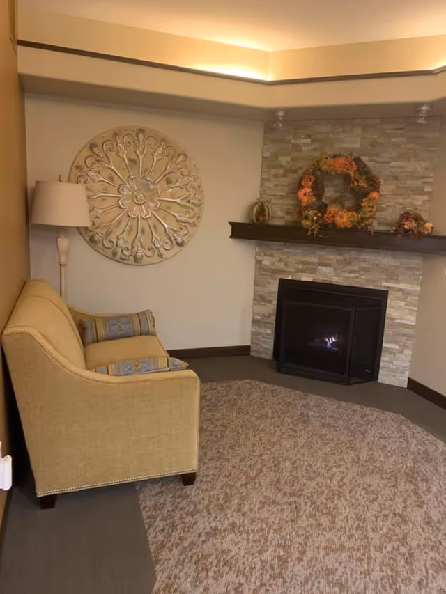 A cozy living room area with a beige upholstered loveseat, a floor lamp, and a decorative round wall art piece. The room features a stone fireplace with a wooden mantel adorned with autumn-themed wreaths and decorations. The floor is covered with a patterned carpet.