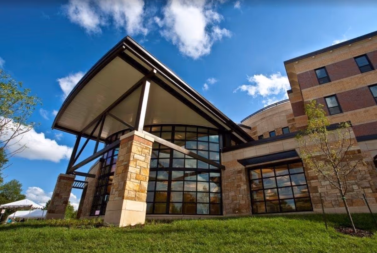 Exterior view of a modern building with large glass windows, stone pillars, and a slanted roof under a blue sky with scattered clouds. There is green grass and a small tree in the foreground.