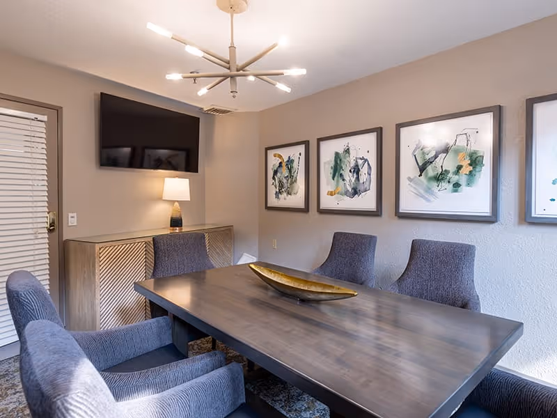 A modern dining room with a rectangular wooden table surrounded by six upholstered gray chairs. On the table is a decorative elongated gold bowl. The walls are beige and decorated with four abstract art paintings in gray frames. A flat-screen TV is mounted on the wall above a wooden sideboard with a lamp on it. A contemporary chandelier with multiple light bulbs hangs from the ceiling.