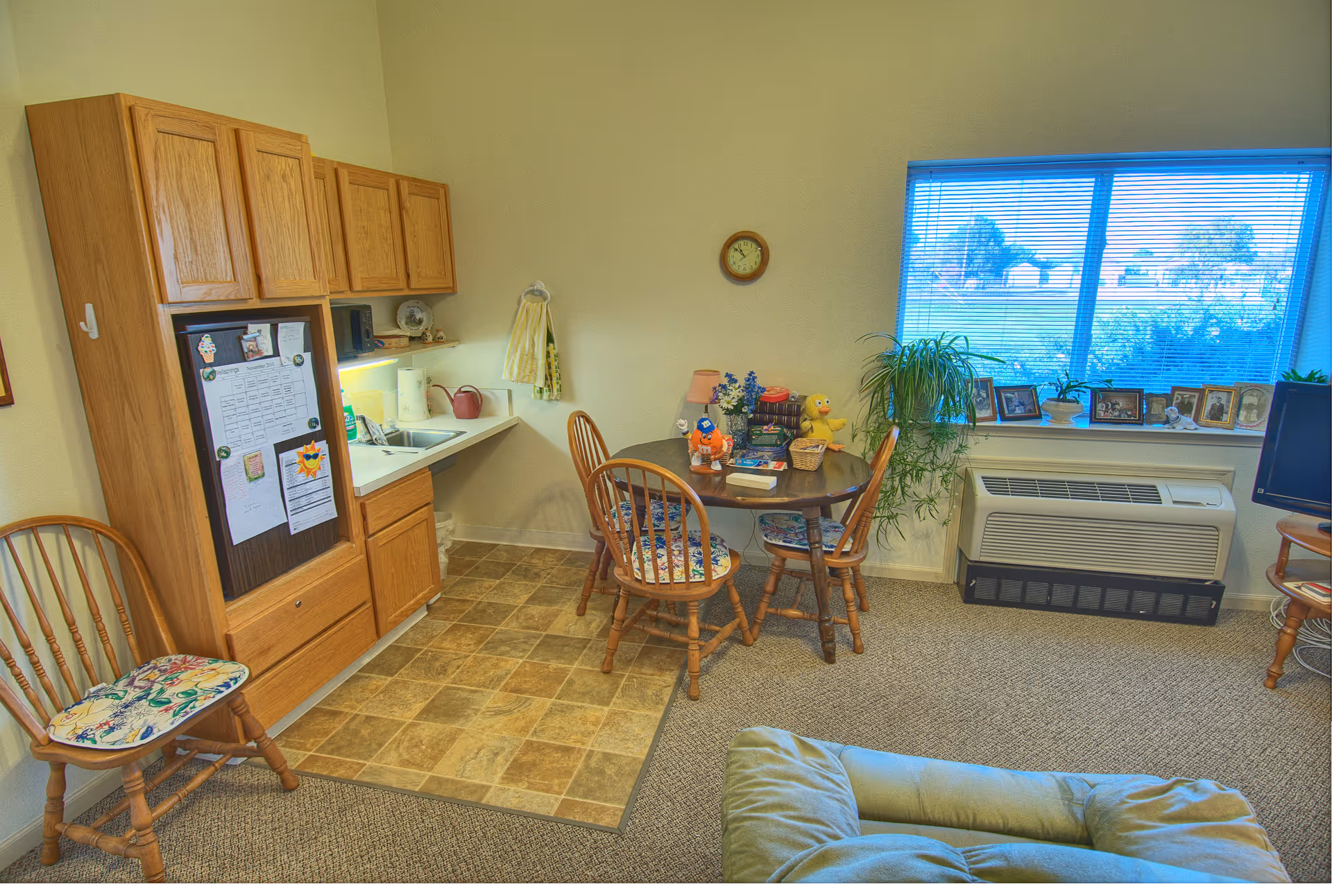 Cozy studio living area with a small kitchenette, round dining table and chairs, a window with plants and framed photos, and a cushioned chair in the foreground.