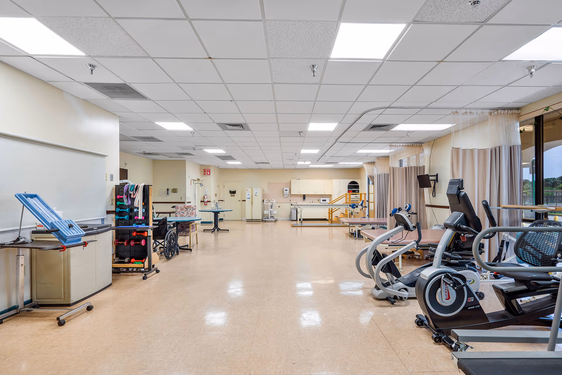 A spacious rehabilitation therapy room with exercise equipment including stationary bikes, parallel bars, and therapy tables. The room has a tiled floor, a drop ceiling with fluorescent lighting, and large windows with curtains on the right side. There are also wheelchairs, weights, and other physical therapy tools visible.