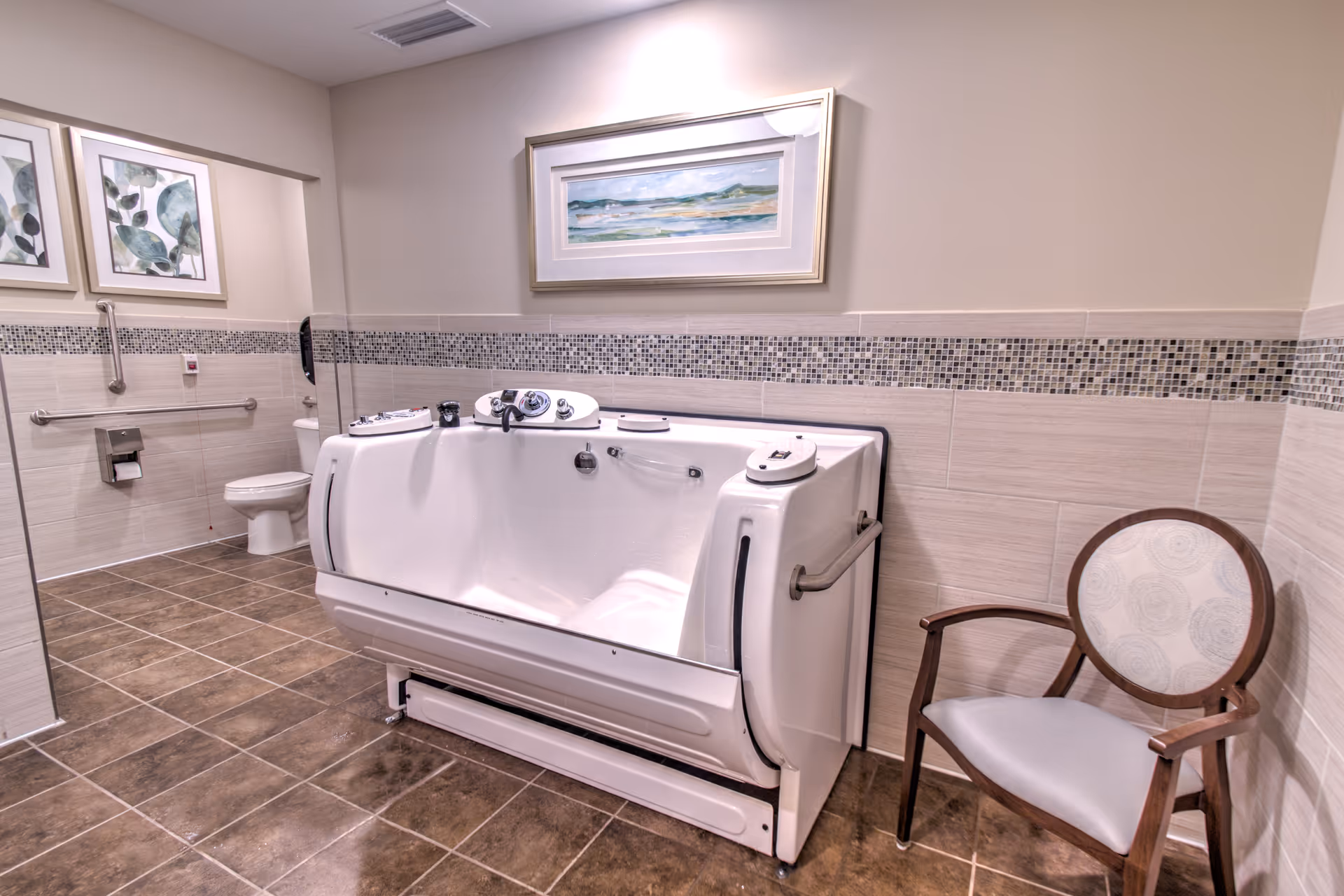 A spacious bathroom with a large walk-in bathtub equipped with handrails and controls, a toilet with grab bars, a wooden chair with cushioned seat and backrest, tiled floor and walls with a decorative mosaic tile border, and framed artwork on the walls.