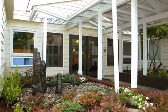 Outdoor garden area at Mckenzie Manor Memory Care featuring a covered walkway with white pillars, various plants, flowers, and decorative rocks.