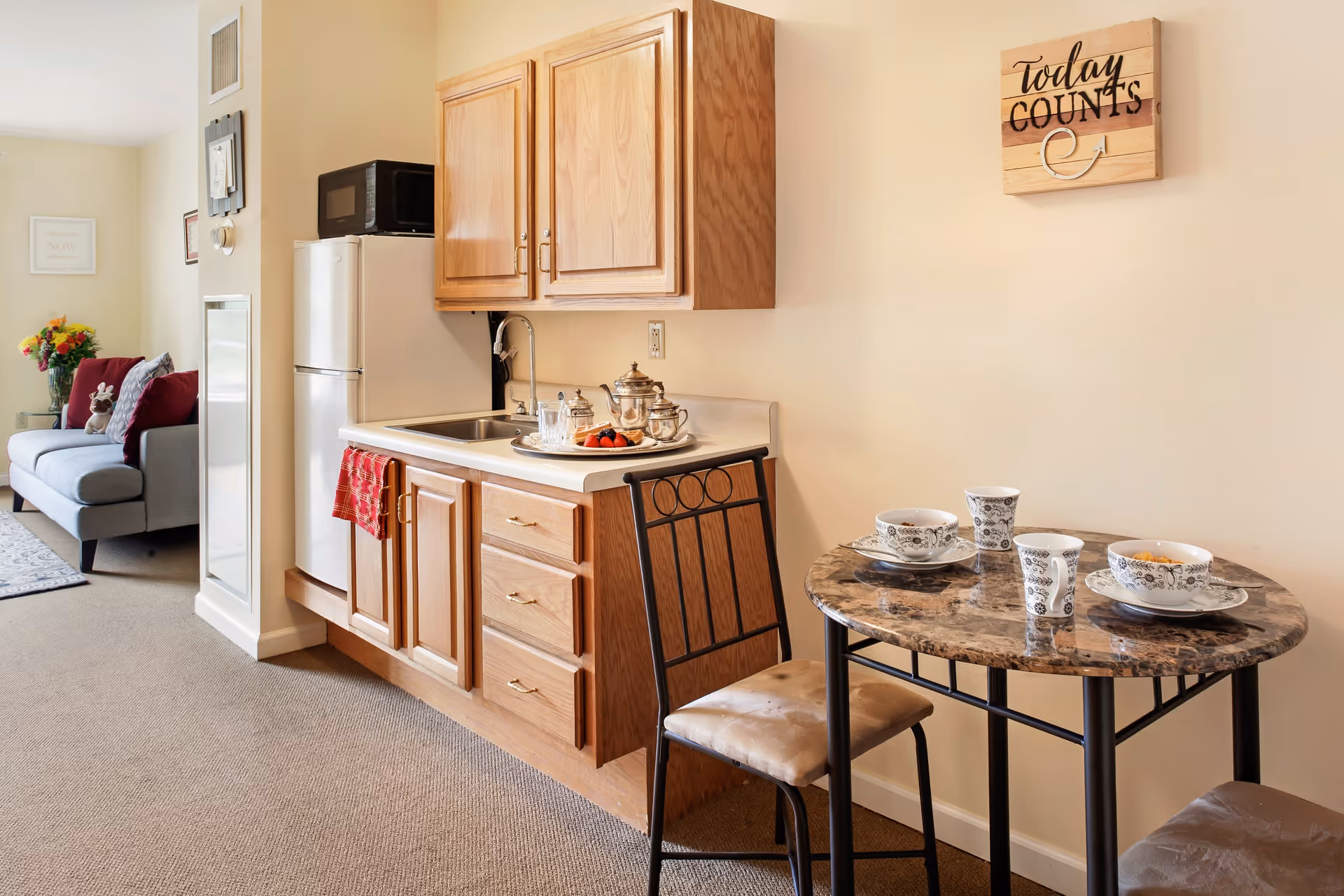Small kitchenette and dining area with wooden cabinets, a mini fridge and sink, a round table set with cups, and a sofa visible in the background.