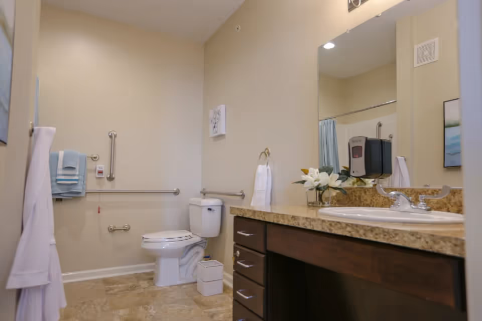 A clean and accessible bathroom with beige walls and tiled floor. The room features a white toilet with grab bars on the walls for support, a granite countertop with a sink, a large mirror above the sink, a soap dispenser mounted on the wall, and neatly folded towels hanging on a towel rack. There is also a small trash bin on the floor and a white hand towel hanging on a ring near the sink.