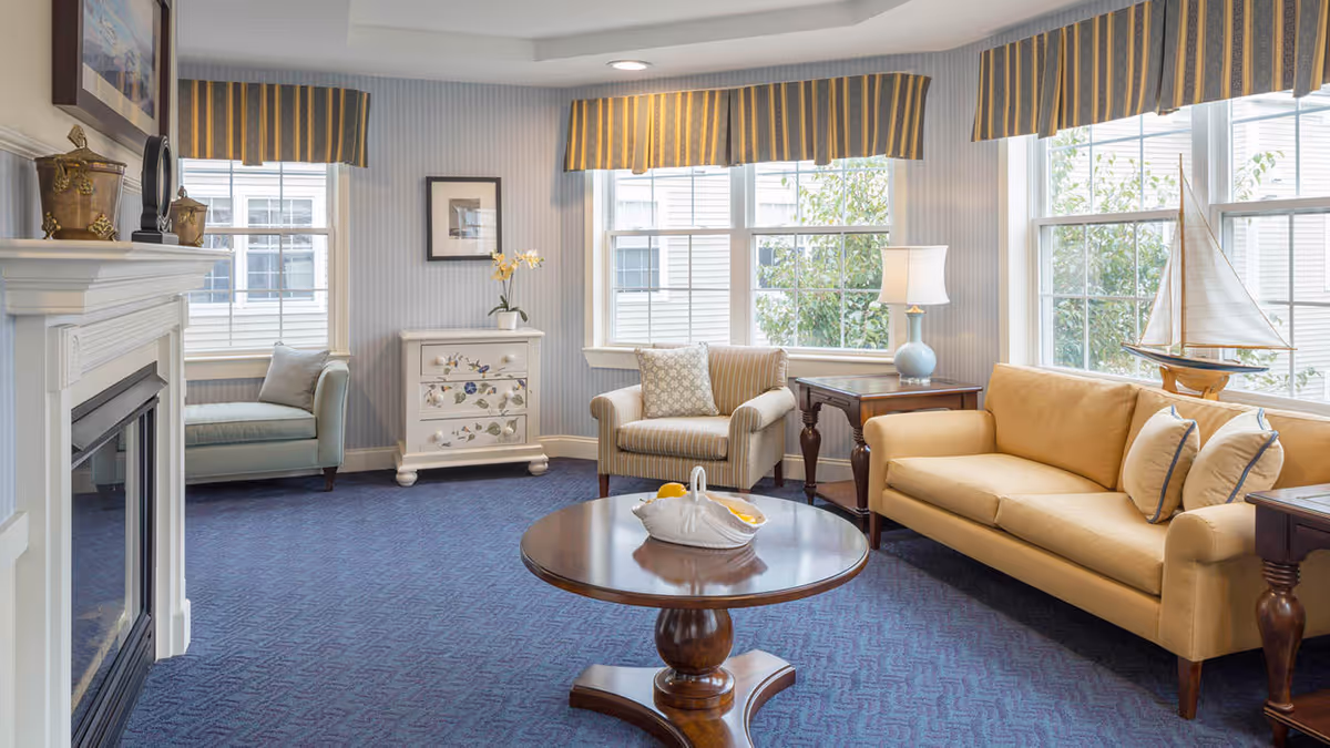 A bright and cozy living room with a yellow sofa, a striped armchair, a small blue loveseat, and a round wooden coffee table with a decorative basket on it. The room has large windows with striped valances, a white fireplace on the left, a small white chest with floral designs, and a model sailboat on a side table. A table lamp is also visible near the sofa.