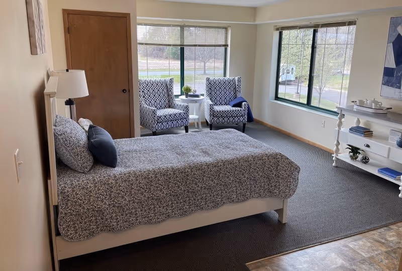A bedroom with a bed covered in a patterned blue and white bedspread, two matching pillows, and a dark blue accent pillow. There is a wooden door on the left wall, a floor lamp beside the bed, and two patterned armchairs with a small white round table between them near large windows showing an outdoor view. On the right side, there is a white console table with decorative items and books, and a large abstract painting on the wall above it.