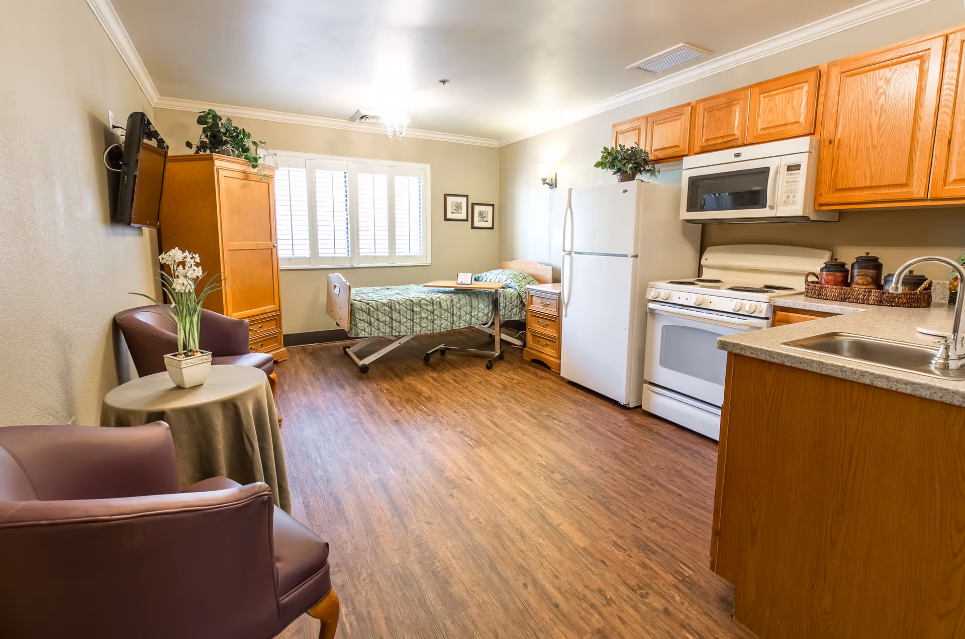 A bright and clean room in a senior living facility featuring a single bed with green patterned bedding near a window with white shutters. The room includes a small kitchen area with wooden cabinets, a white refrigerator, stove, microwave, and a sink. There are two purple armchairs around a small round table with a potted plant, a wooden cabinet with a TV mounted above it, and framed artwork on the wall.