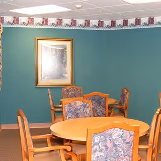 A room with green walls featuring a framed picture and a decorative border near the ceiling. The room contains two wooden dining tables, one round and one rectangular, each surrounded by chairs with floral-patterned upholstery.