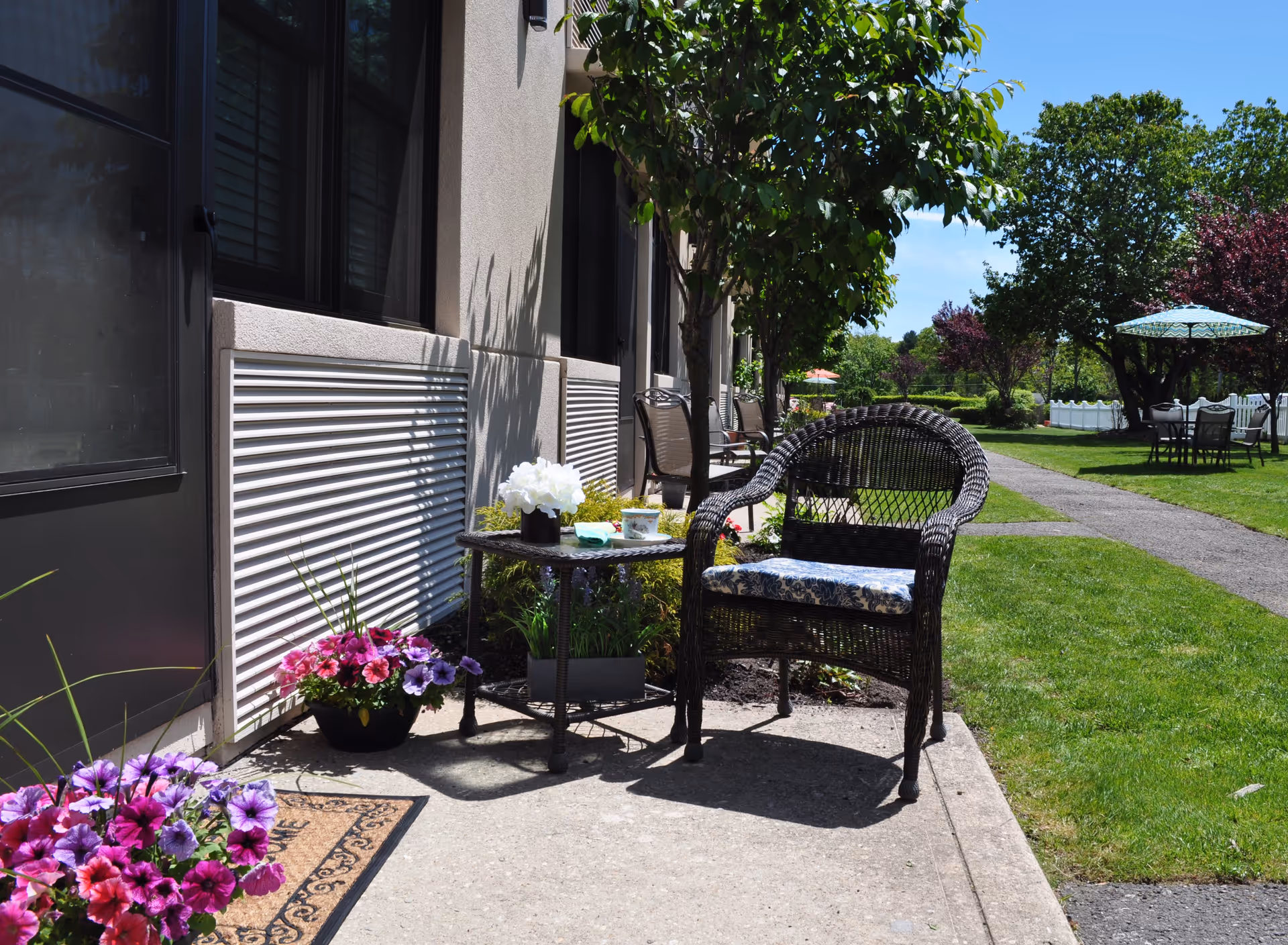 Wicker chair and small table with flowers on a patio outside a senior living building overlooking a grassy courtyard with tables and umbrellas.