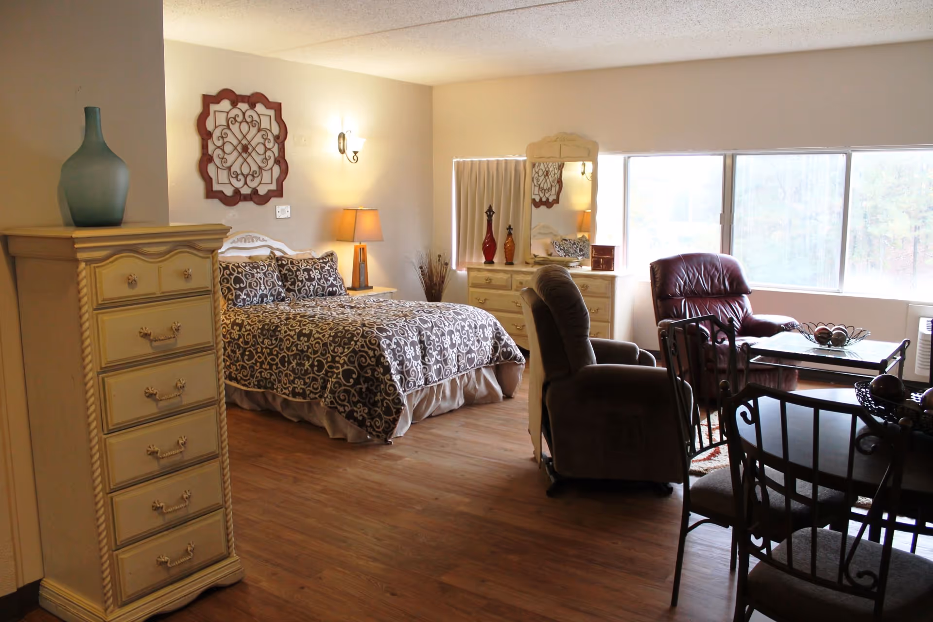 A cozy senior living facility room with a bed covered in patterned bedding, a wooden dresser with a large blue vase on top, a nightstand with a lamp, a dresser with a mirror and decorative vases, two armchairs, a small table, and a dining table with chairs near a large window letting in natural light.