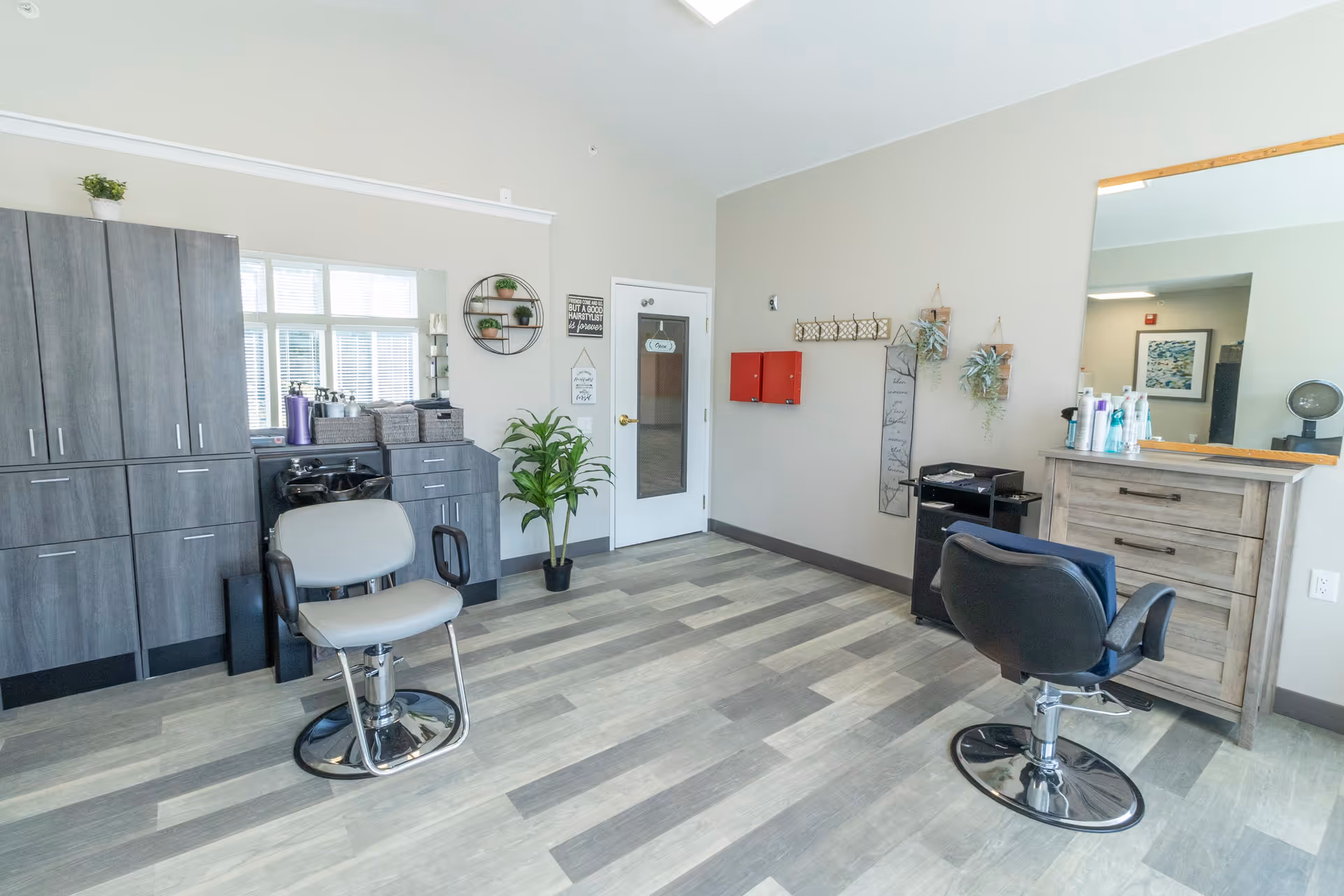 A bright and clean hair salon area inside a senior living facility with two salon chairs, a large mirror, wooden cabinets, a plant, and various hair care products on the counter. The room has light-colored walls and flooring with natural light coming through a window.