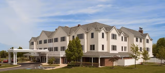 Large multi-story beige assisted living building with a covered entrance, lawn and trees under a blue sky.