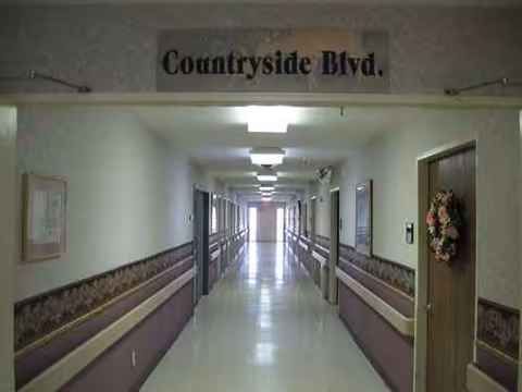 Long, well-lit corridor in a senior living facility with doors, rails, and a wreath.