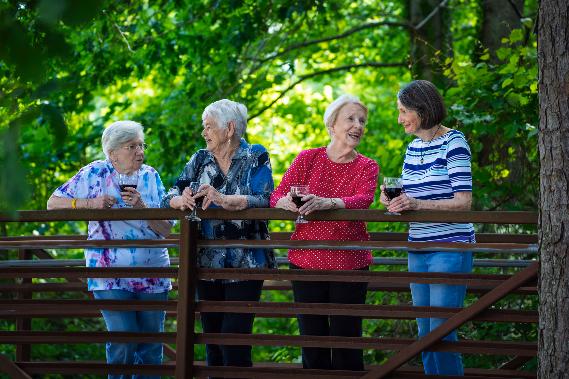 Four elderly women standing on a wooden bridge outdoors surrounded by green trees, each holding a glass of red wine and engaging in conversation.