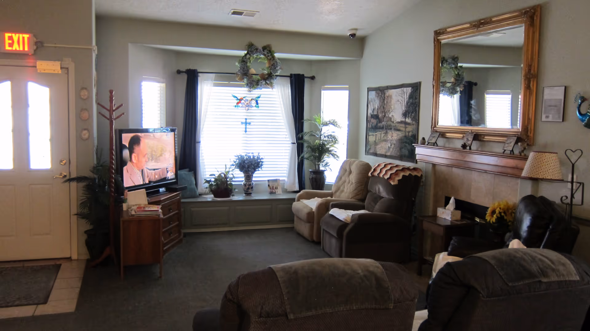 A cozy living room with multiple armchairs arranged around a fireplace with a large ornate mirror above it. A TV is on a wooden stand near a window with white blinds and dark curtains. Various plants and decorative items are placed around the room, and an exit sign is visible above a door on the left.