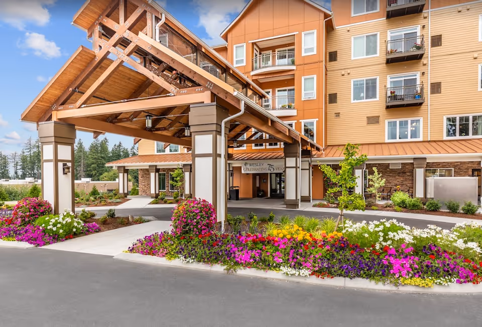 Entrance of Wesley Homes Bradley Park facility featuring a large covered driveway with wooden beams, surrounded by colorful flower beds and landscaping. The multi-story building has balconies with flower pots and a sign that reads 'Wesley Celebrating 75 Years'.