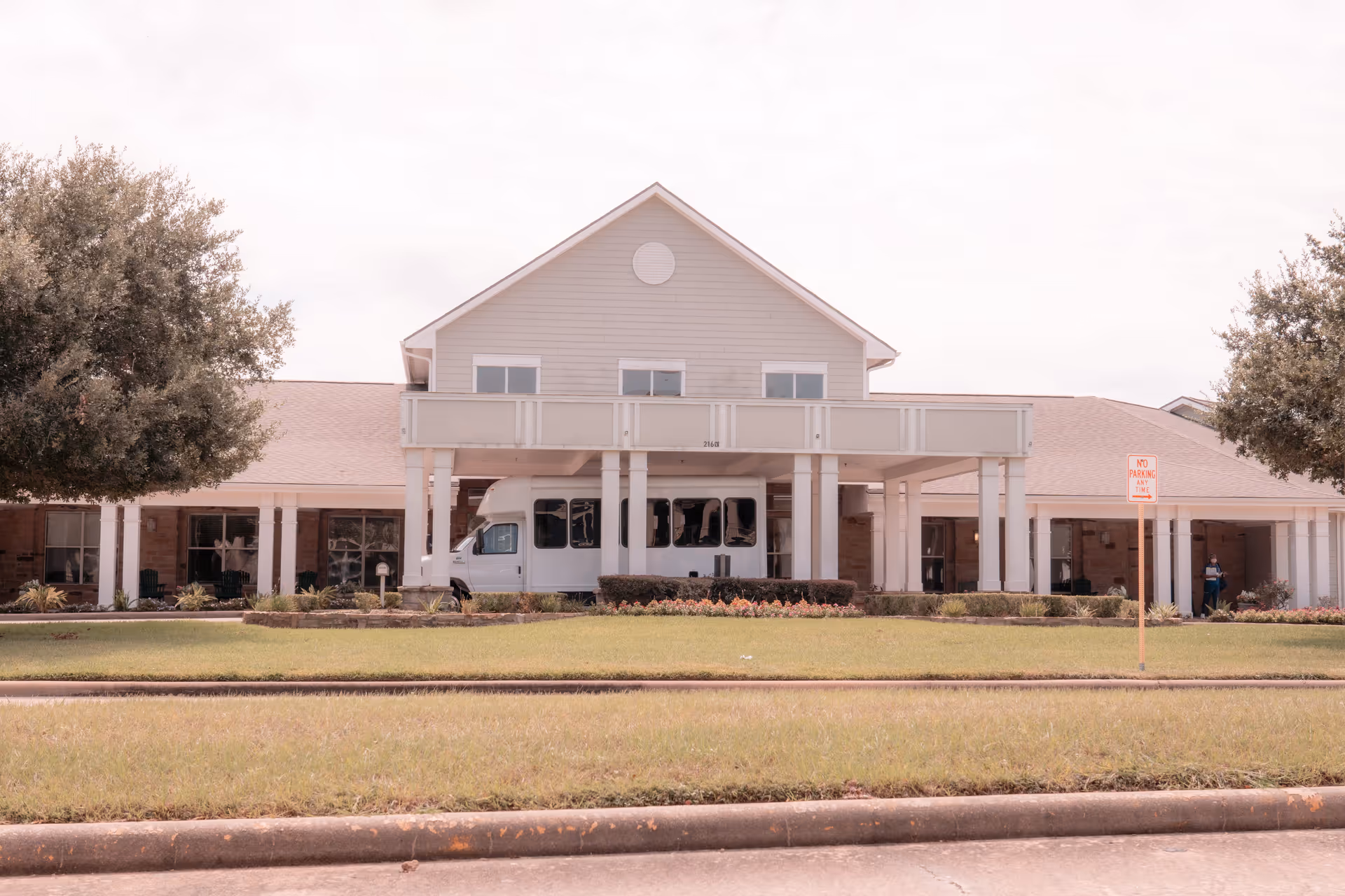 Front exterior of an assisted living facility with a covered entrance, parked shuttle van, and landscaped lawn.